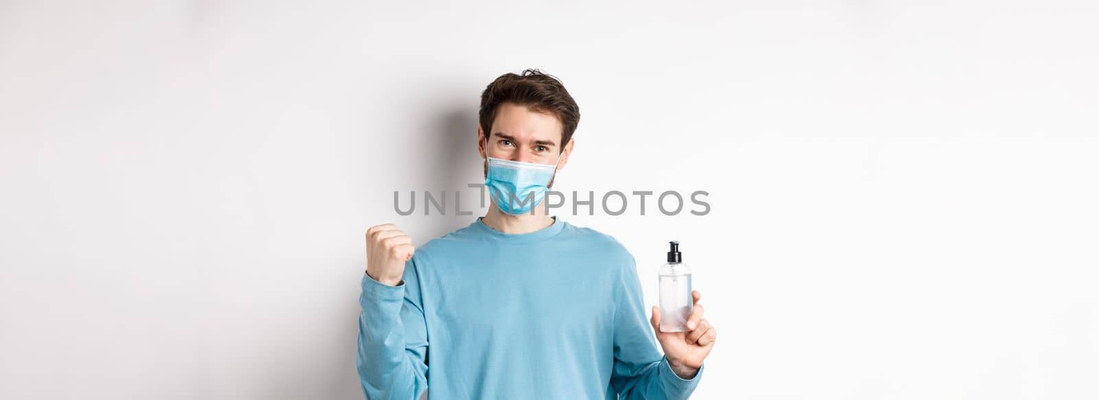 Covid-19, health and quarantine concept. Cheerful man in face mask celebrating, showing fist pump and bottle with hand sanitizer, fighting germs, white background.