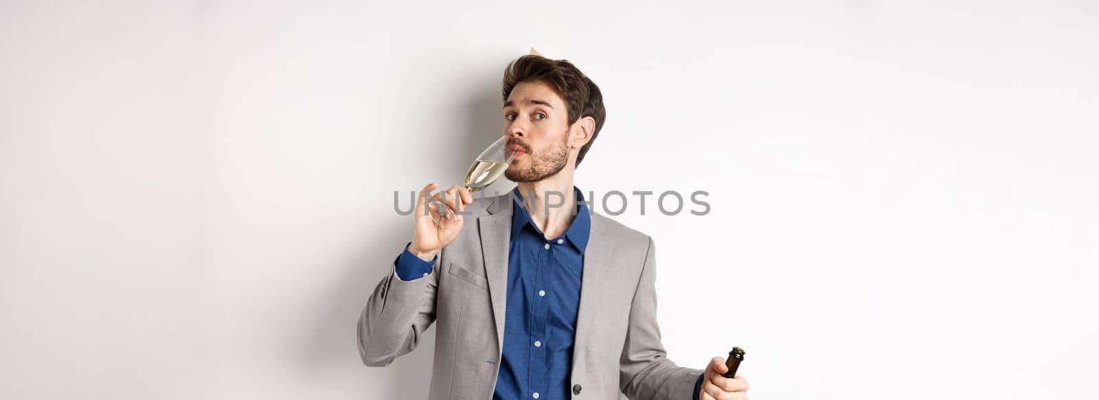 Celebration and holidays concept. Handsome bearded man in suit and birthday hat holding bottle, drinking glass of champagne, standing on white background.