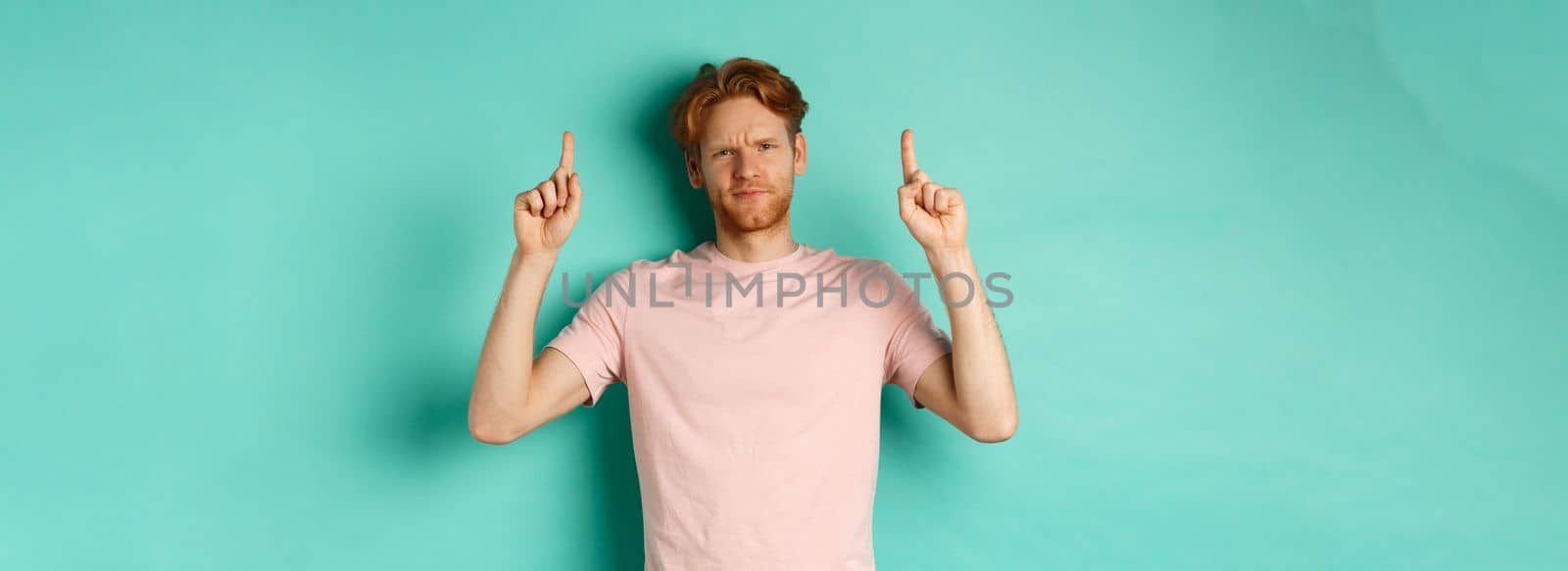 Skeptical and disappointed redhead man pointing fingers up, frowning displeased and looking upset, standing over turquoise background by Benzoix