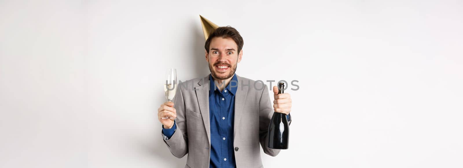 Celebration and holidays concept. Excited handsome birthday guy in party hat smiling, drinking champagne and having fun, white background.