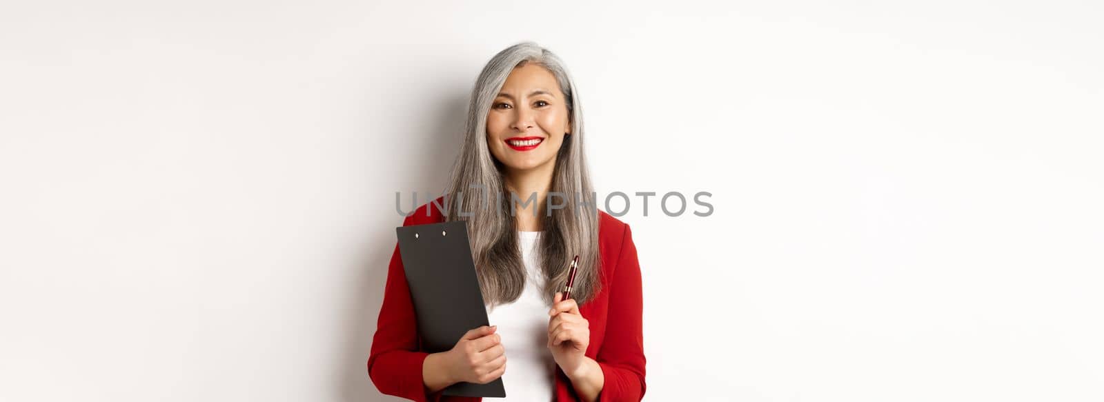 Successful asian lady boss in red blazer, holding clipboard with documens and pen, working and looking happy, white background.