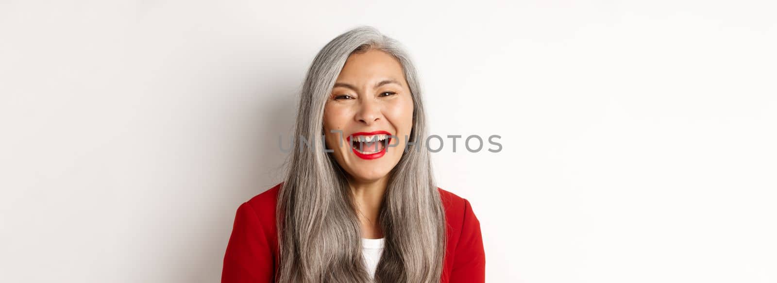 Close-up of happy asian businesswoman with long grey hair, wearing red blazer, laughing and smiling joyfully at camera, white background by Benzoix