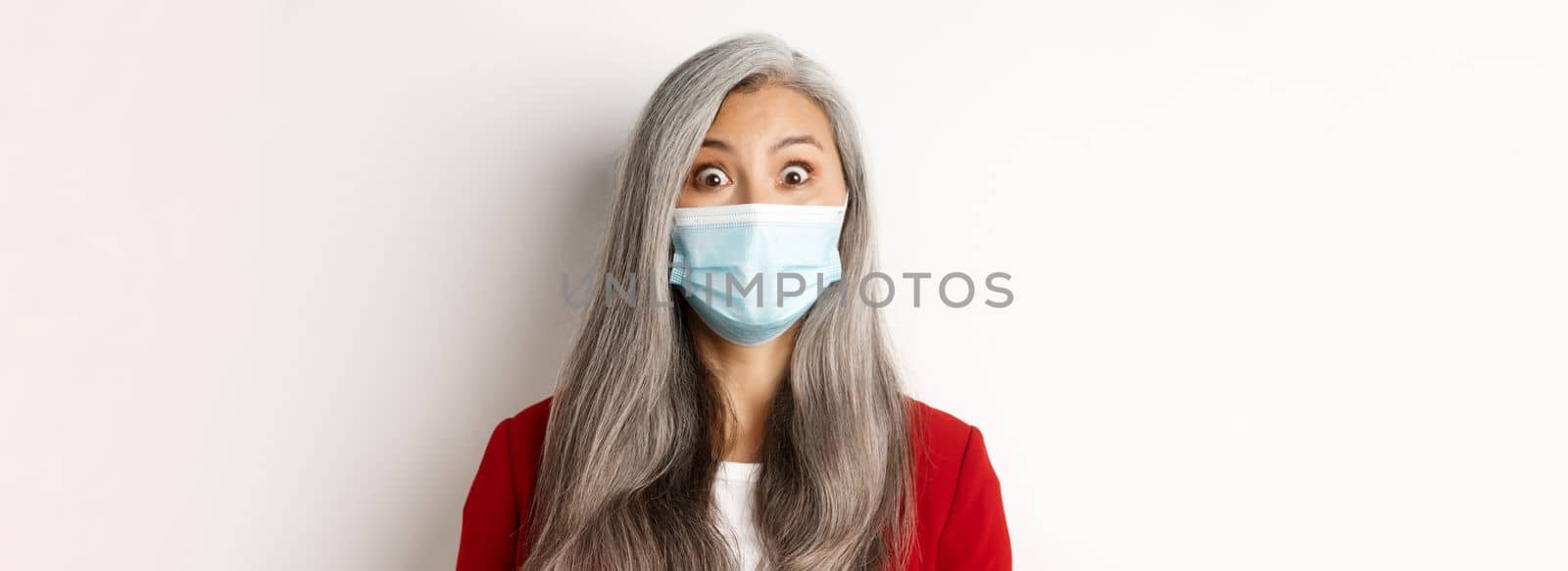 Covid, pandemic and business concept. Close up of surprised asian grandmother in medical mask raising eyebrows, staring amazed at camera, white background.
