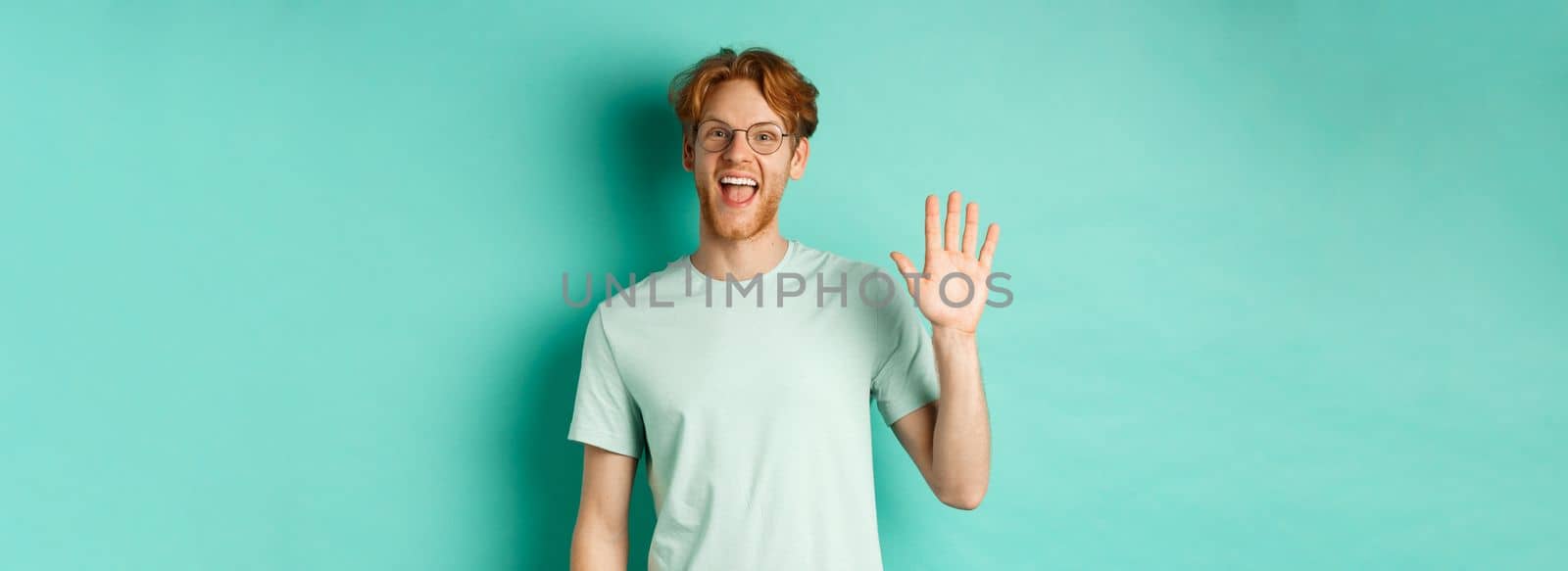 Friendly bearded guy in glasses saying hello, waving hand to greet and welcome you, standing cheerful and smiling over turquoise background by Benzoix