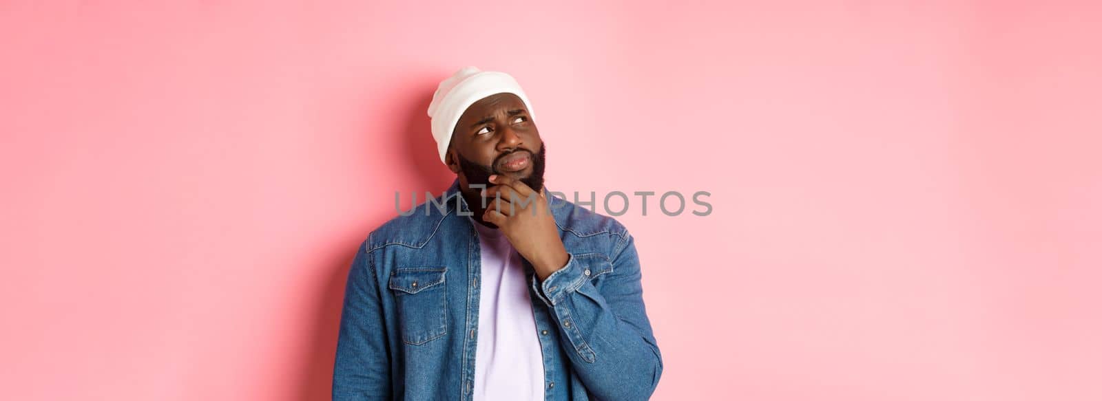 Image of puzzled and concerned black man, staring at upper left corner and thinking, making decision with doubts, standing over pink background.