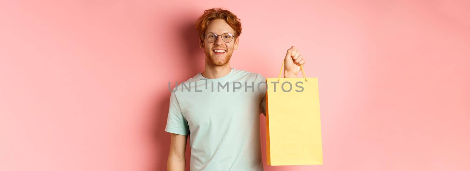 Handsome young man buying presents, holding shopping bag and smiling at camera, standing over pink background by Benzoix
