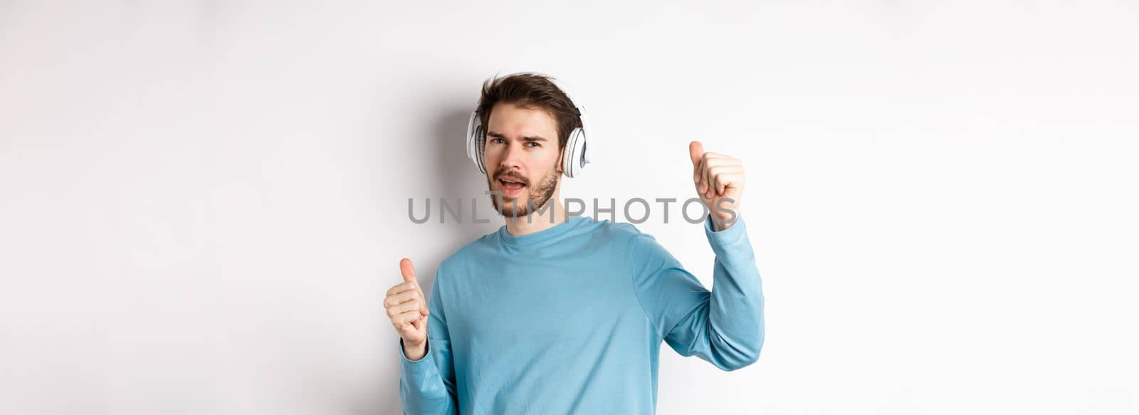 Cheeky guy dancing and having fun in wireless headphones, enjoying good sound quality, white background by Benzoix