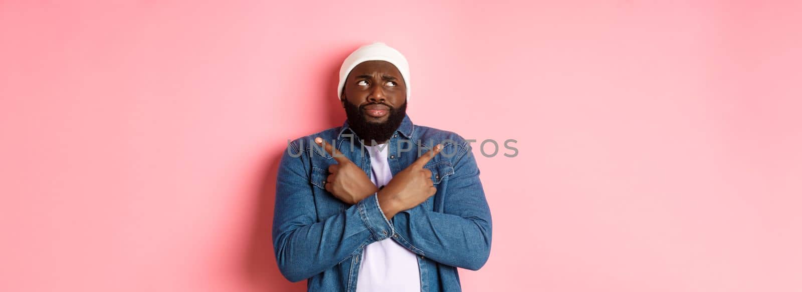 Confused Black man with beard, making choice, pointing fingers sideways and looking puzzled, standing over pink background.