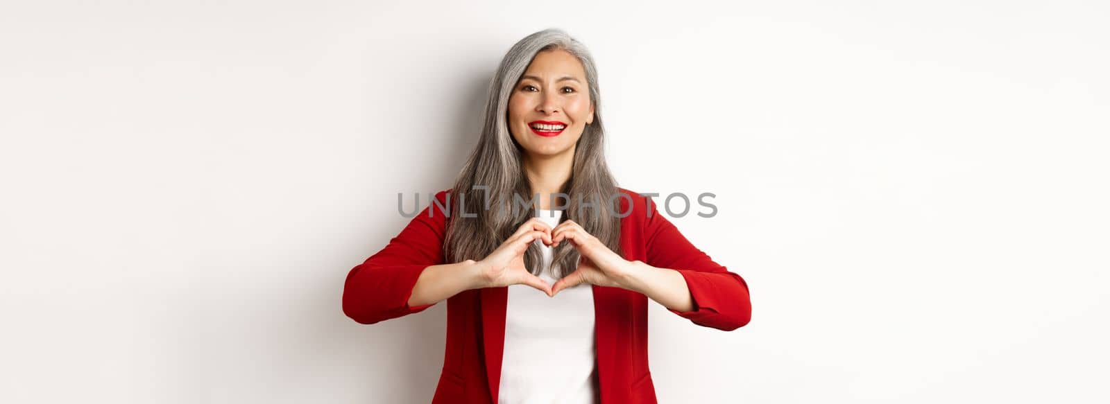 Beautiful asian mature woman in red blazer and makeup, showing heart sign and smiling, I love you gesture, standing over white background.