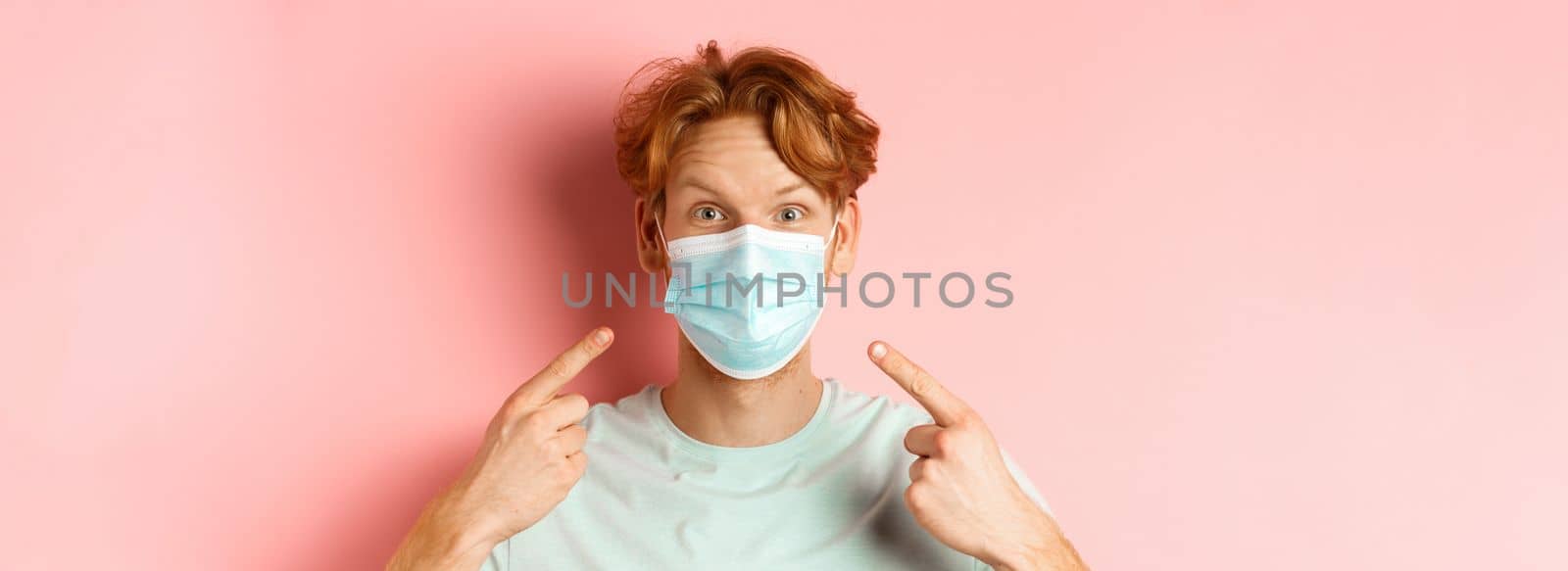 Covid-19 and pandemic concept. Close up of redhead man with messy hairstyle, pointing fingers at medical mask from coronavirus, standing over pink background by Benzoix