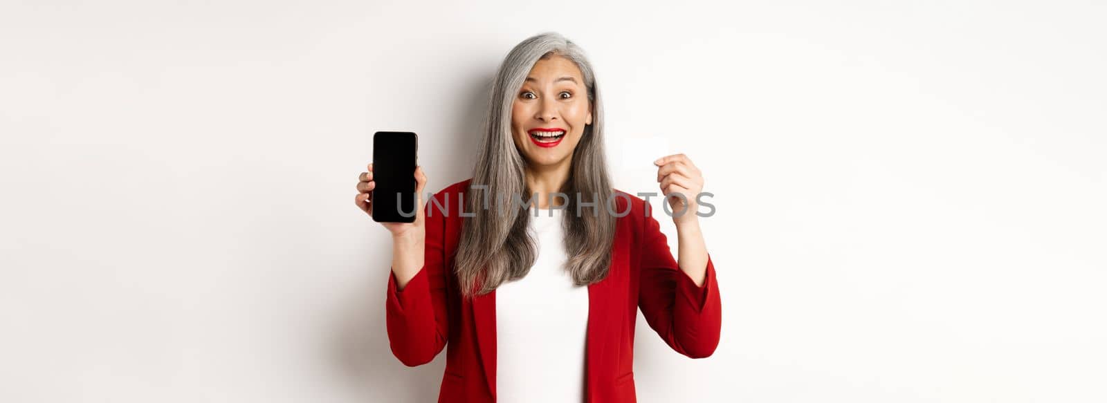Cheerful asian mature woman showing blank smartphone screen and credit card, concept of e-commerce.