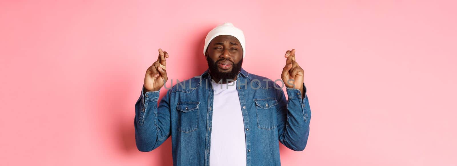 Image of young african-american man whining, pleading god, cross fingers and crying, standing over pink background.