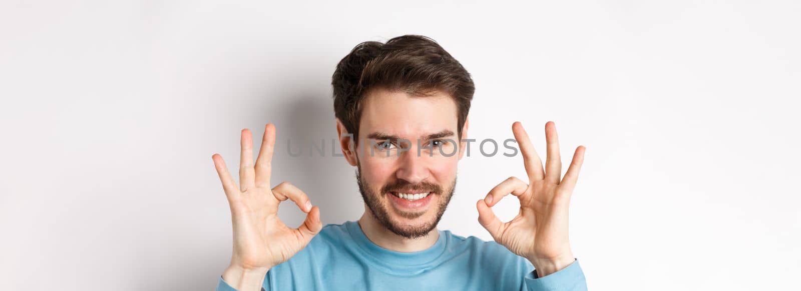 Close-up of confident young man smiling, showing okay signs, praising excellent choice, approve something good, standing over white background.