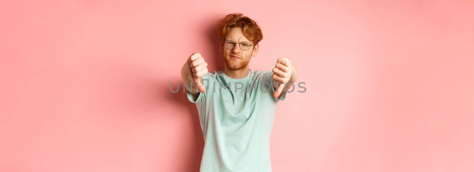 Disappointed young man in glasses, with messy red haircut, showing thumbs down and grimacing unsatisfied, express dislike, standing over pink background.