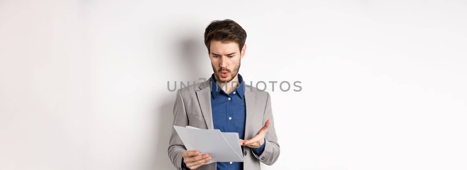 Confused businessman looking at bad document, complaining on deal, pointing at paper frustrated, standing in suit on white background.