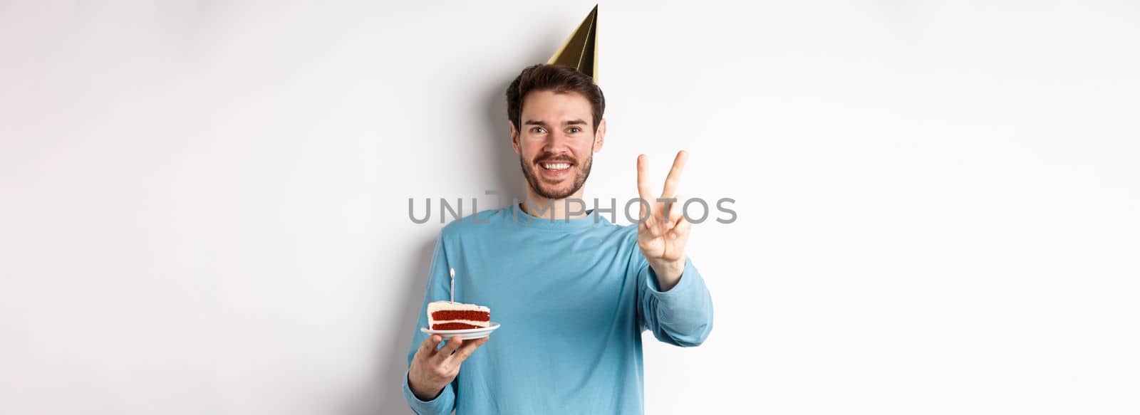 Celebration and holidays concept. Happy young man celebrating birthday, taking picture with peace sign, wearing party hat and holding bday cake, white background by Benzoix