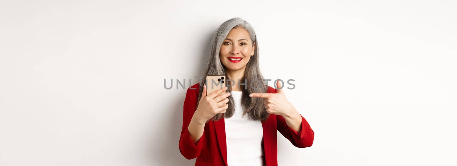 Asian elderly woman in elegant blazer showing smartphone, pointing finger at mobile phone and smiling, standing over white background by Benzoix