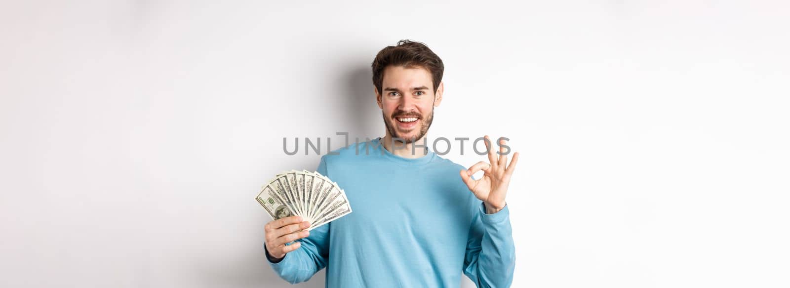 Handsome young man showing quick loans money, make okay gesture and smiling with cash, standing over white background by Benzoix