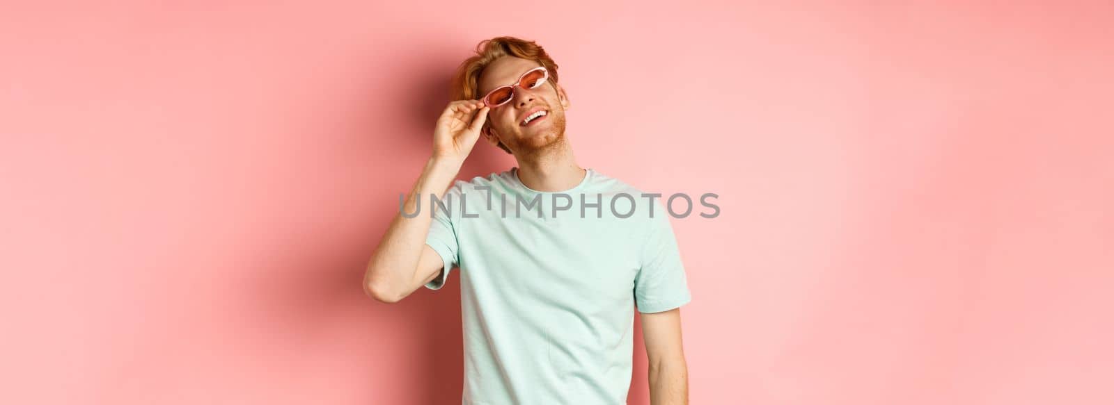 Tourism and vacation concept. Happy young man with red hair relaxing, wearing sunglasses and smiling carefree, standing over pink background.