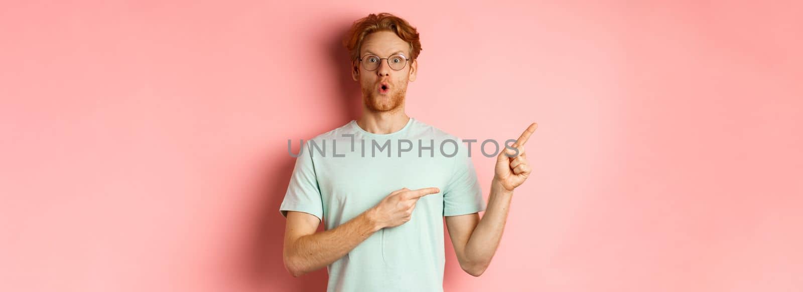 Shopping concept. Portrait of man with red hair and beard, wearing glasses with summer t-shirt, pointing fingers at upper left corner and saying wow impressed, pink background.