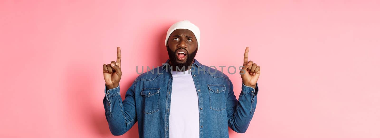 Displeased african american guy in beanie, pointing fingers up and staring at camera bothered, complaining, standing over pink background.