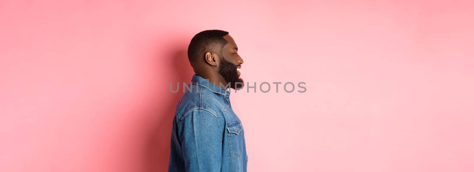 Profile of handsome bearded Black guy standing over pink background, smiling and looking left at copy space by Benzoix