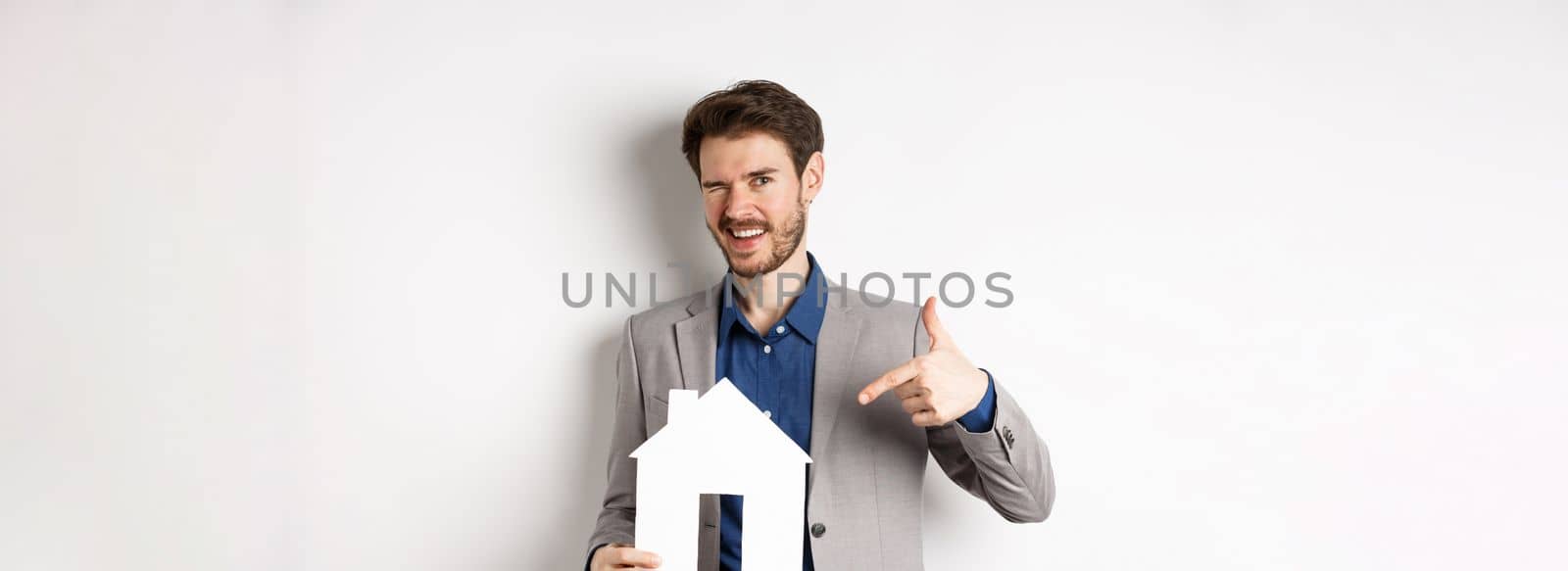 Real estate. Handsome businessman winking and pointing at paper house cutout, advertising company, standing on white background.