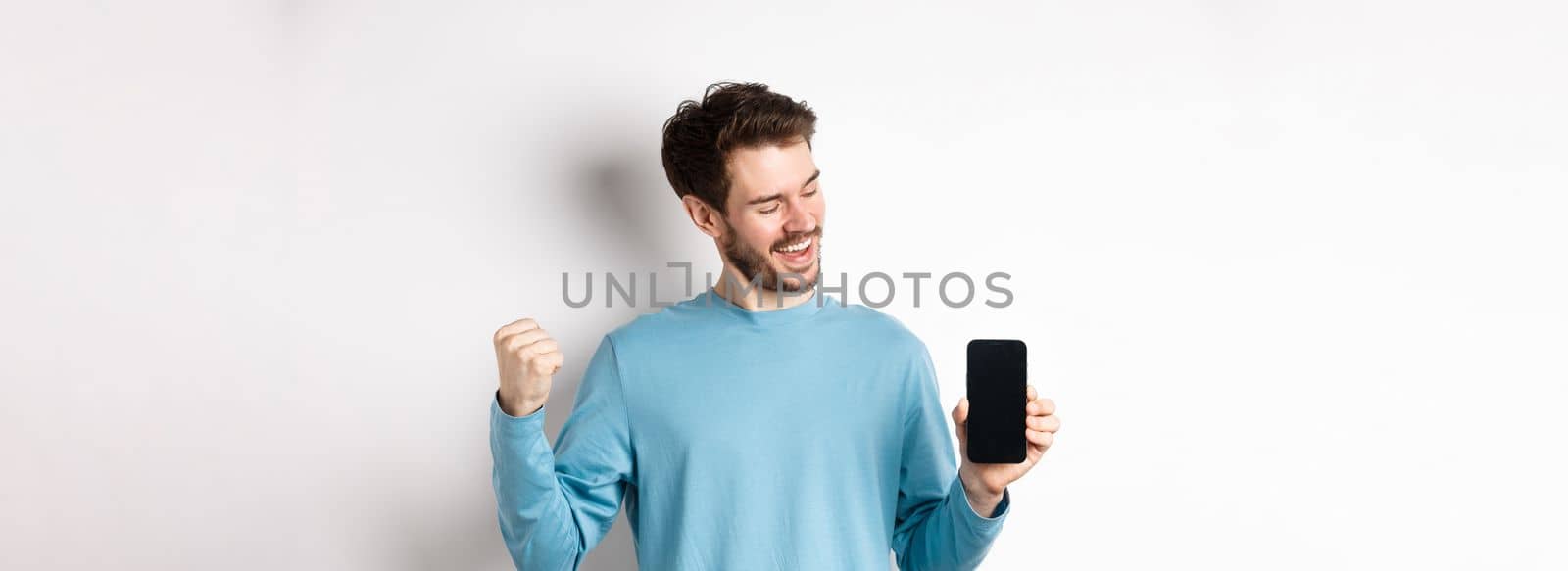 E-commerce and shopping concept. Lucky man winning online, showing empty smartphone screen and triumphing, saying yes with fist pump, standing over white background by Benzoix