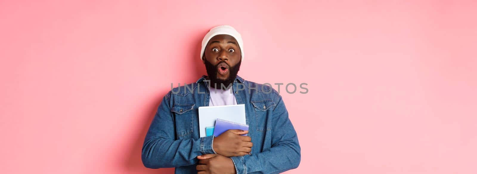 Education. Excited african-american adult student carry notebooks, staring at camera amaze, standing over pink background.
