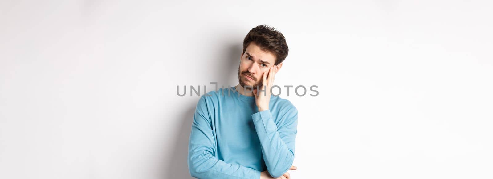 Sad young man looking frustrated, touching temple of head and frowning uneasy, standing against white background.