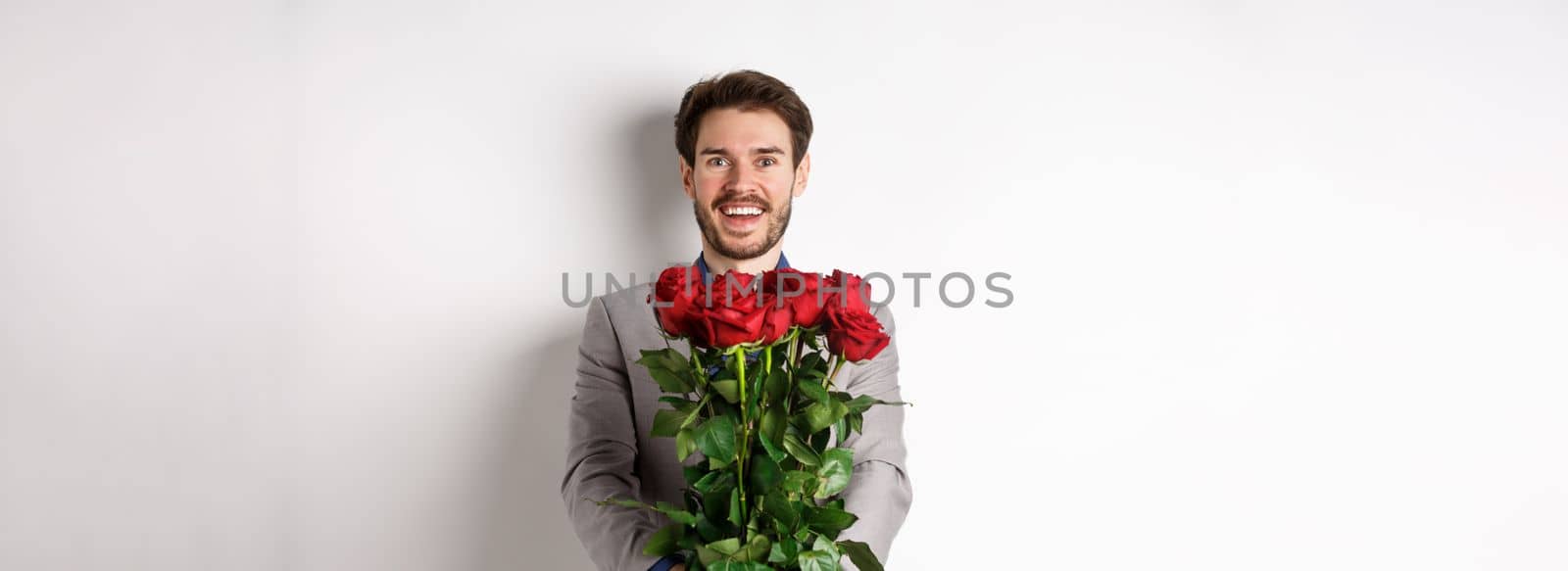 Handsome man in love wishing happy valentines day, giving bouquet of flowers on romantic date, smiling at camera, wearing suit over white background by Benzoix