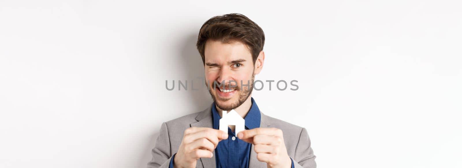 Real estate and insurance concept. Handsome man in suit winking and smiling, showing small paper house cutout, standing on white background.