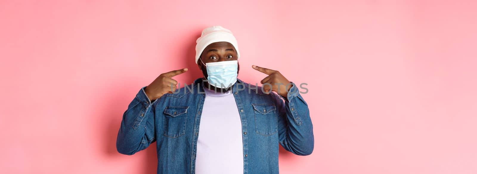 Coronavirus, lifestyle and social distancing concept. Happy Black man in beanie pointing at his face mask, smiling at camera, standing over pink background.