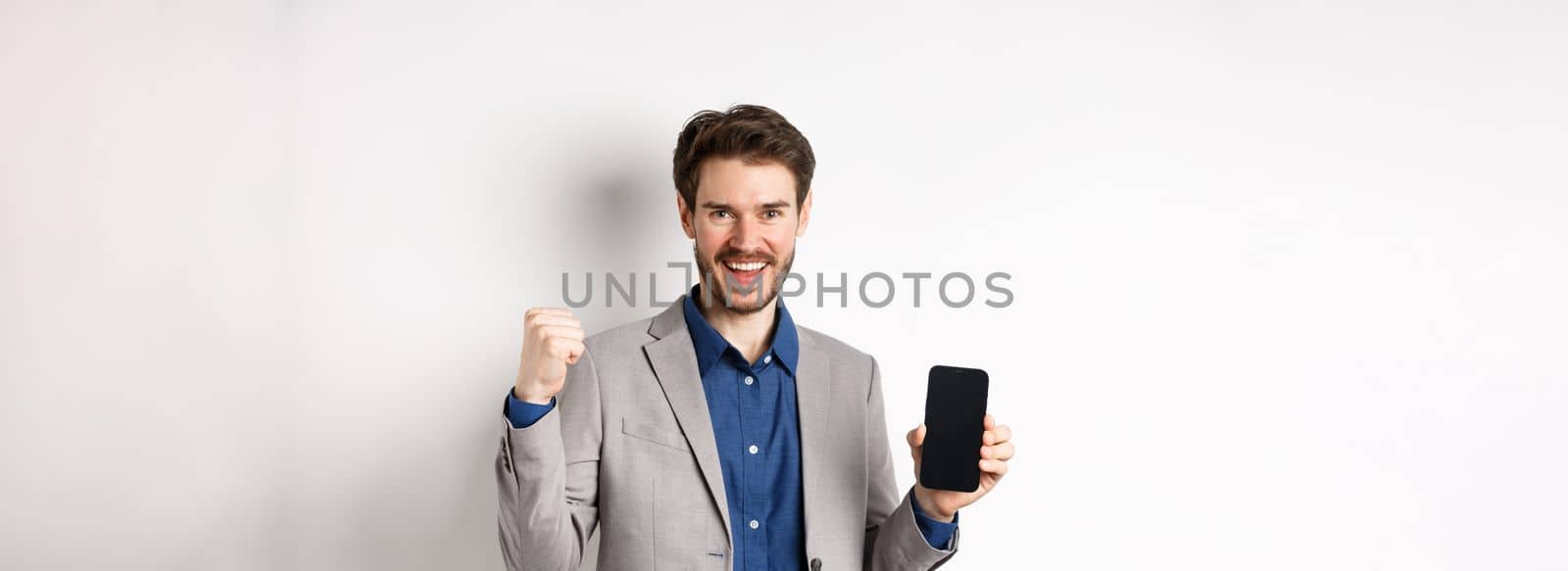 E-commerce and online shopping concept. Man making money in internet, showing smartphone screen and winner gesture, smiling satisfied, standing on white background.