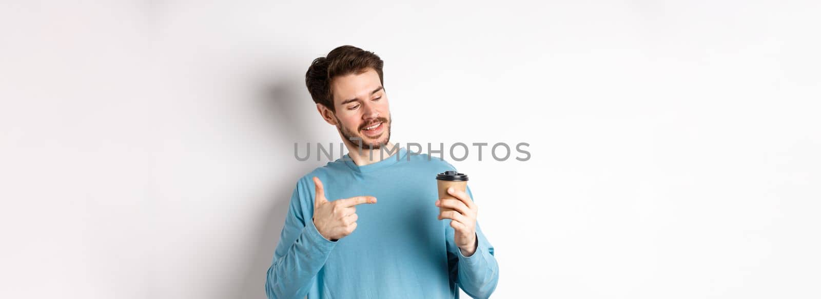 Handsome caucasian man pointing finger at coffee cup, recommending cafe, standing on white background satisfied.