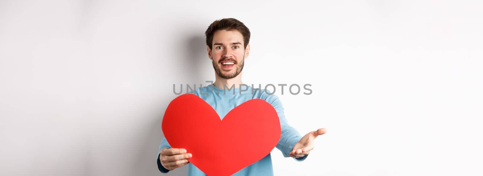 Handsome man in love making confession to you, pointing hand at camera, holding big red heart cutout on valentines day, singing romantic serenade, standing over white background.