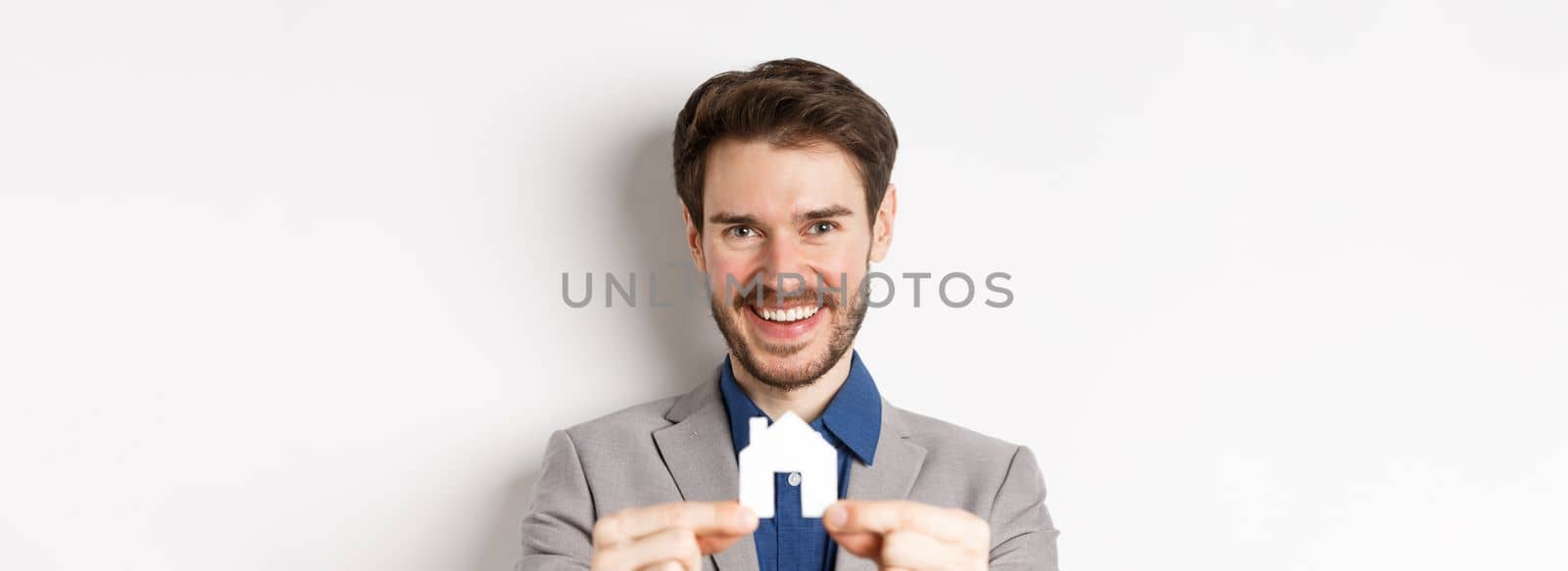 Real estate and insurance concept. Handsome man in suit smiling, showing small paper house cutout, standing on white background.