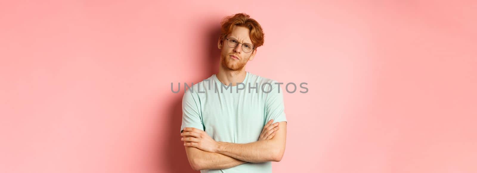 Young man with red hair and beard, wearing glasses and t-shirt, cross arms on chest, frowning while staring with skeptical and doubtful expression, standing over pink background by Benzoix