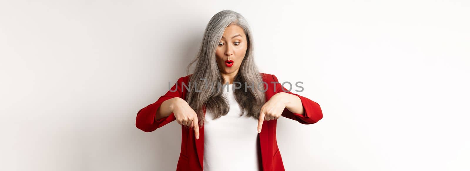 Impressed asian mature woman checking out promo, pointing and looking down with amazed face, standing in blazer over white background.