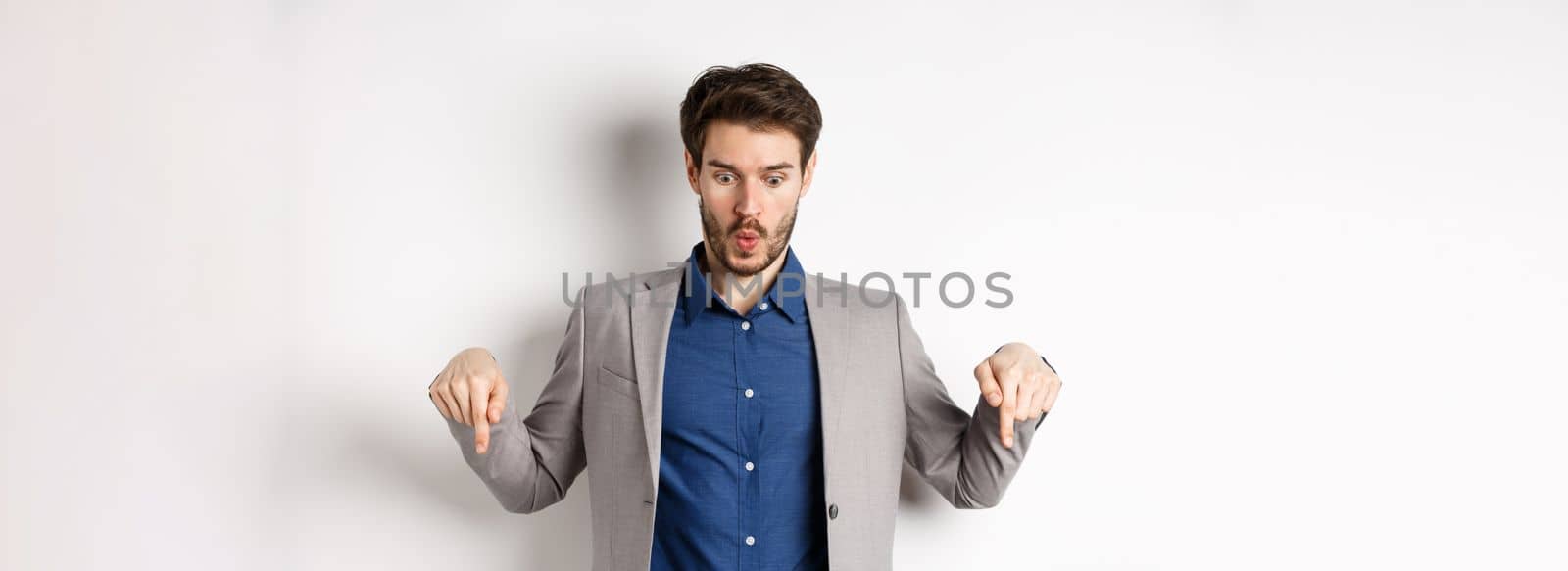 Wow look at this. Impressed and excited businessman in suit pointing fingers down, looking at bottom advertisement with amazed face, standing on white background.