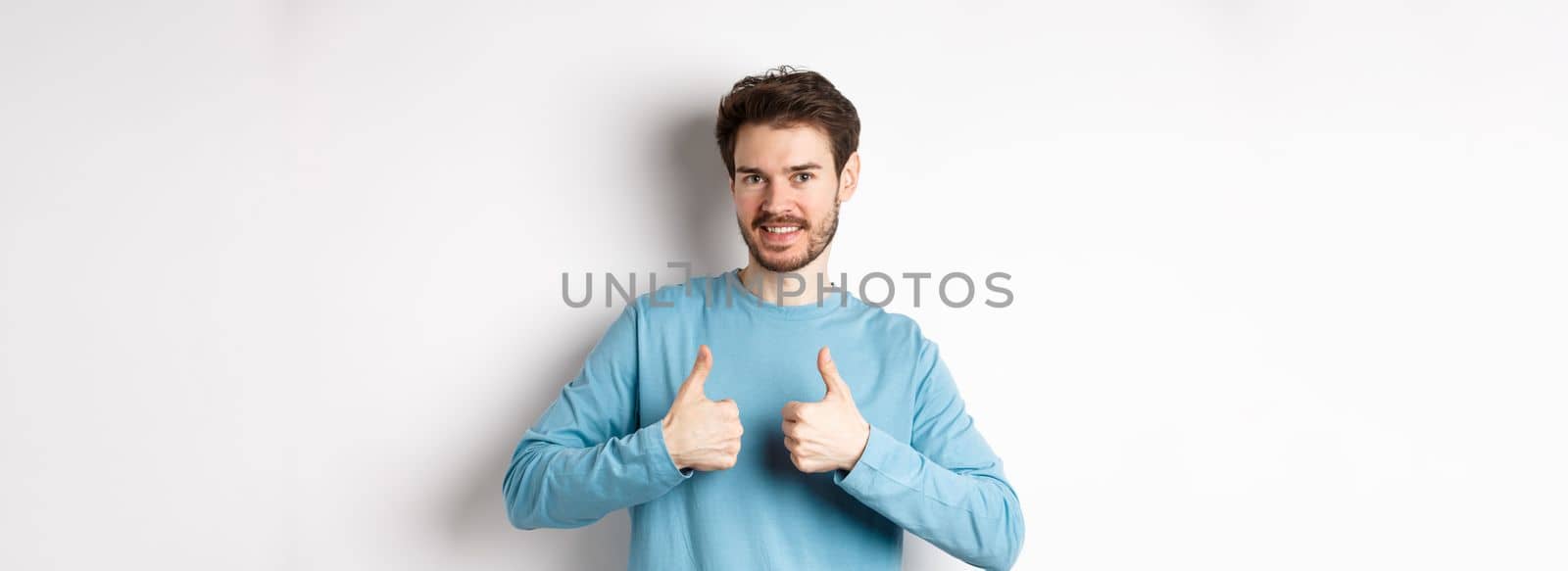 Good choice. Attractive caucasian man with beard showing thumbs up and smiling, like and approve good product, recommending something, standing on white background.