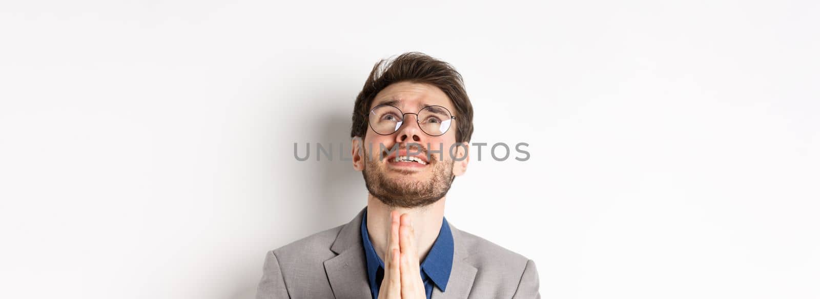 Nervous hopeful man in glasses and suit begging god, asking please and shaking hands in pray, white background.