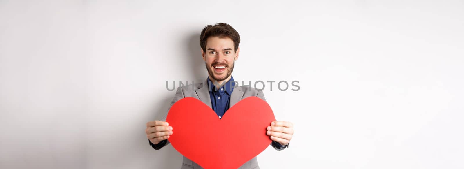 Handsome young man wishing happy Valentines day, giving big red heart sign and smiling, make surprise to lover, standing in suit over white background by Benzoix