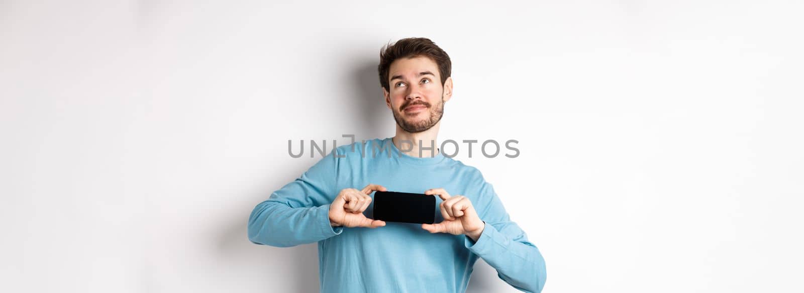 E-commerce and shopping concept. Smiling young man showing mobile screen and dreaming about something, looking at upper right corner nostalgic, standing over white background.