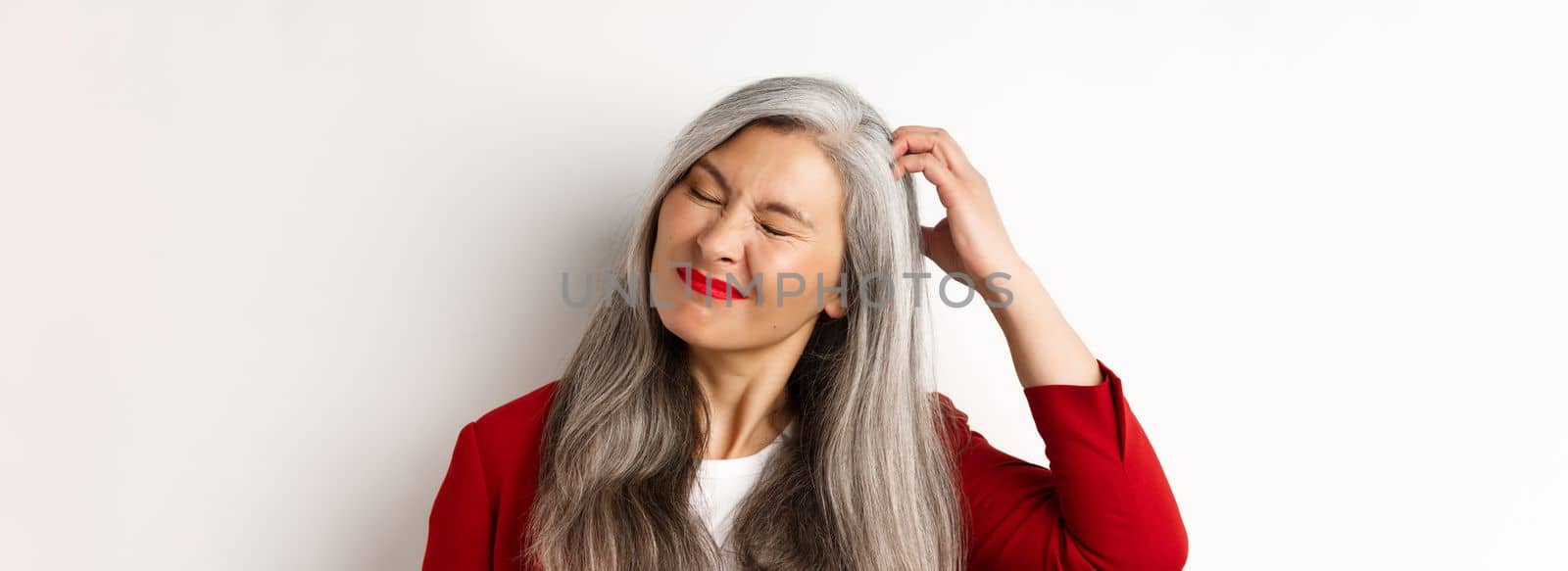 Close up of senior asian woman scratching head and looking bothered by itching, standing over white background.