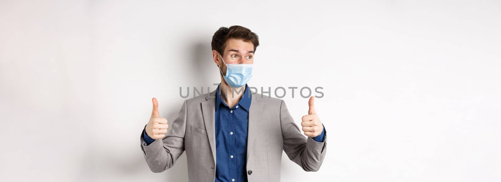 Covid-19, pandemic and business concept. Excited businessman in face mask and suit, showing thumbs up and looking aside at logo impressed, praise good thing, white background.