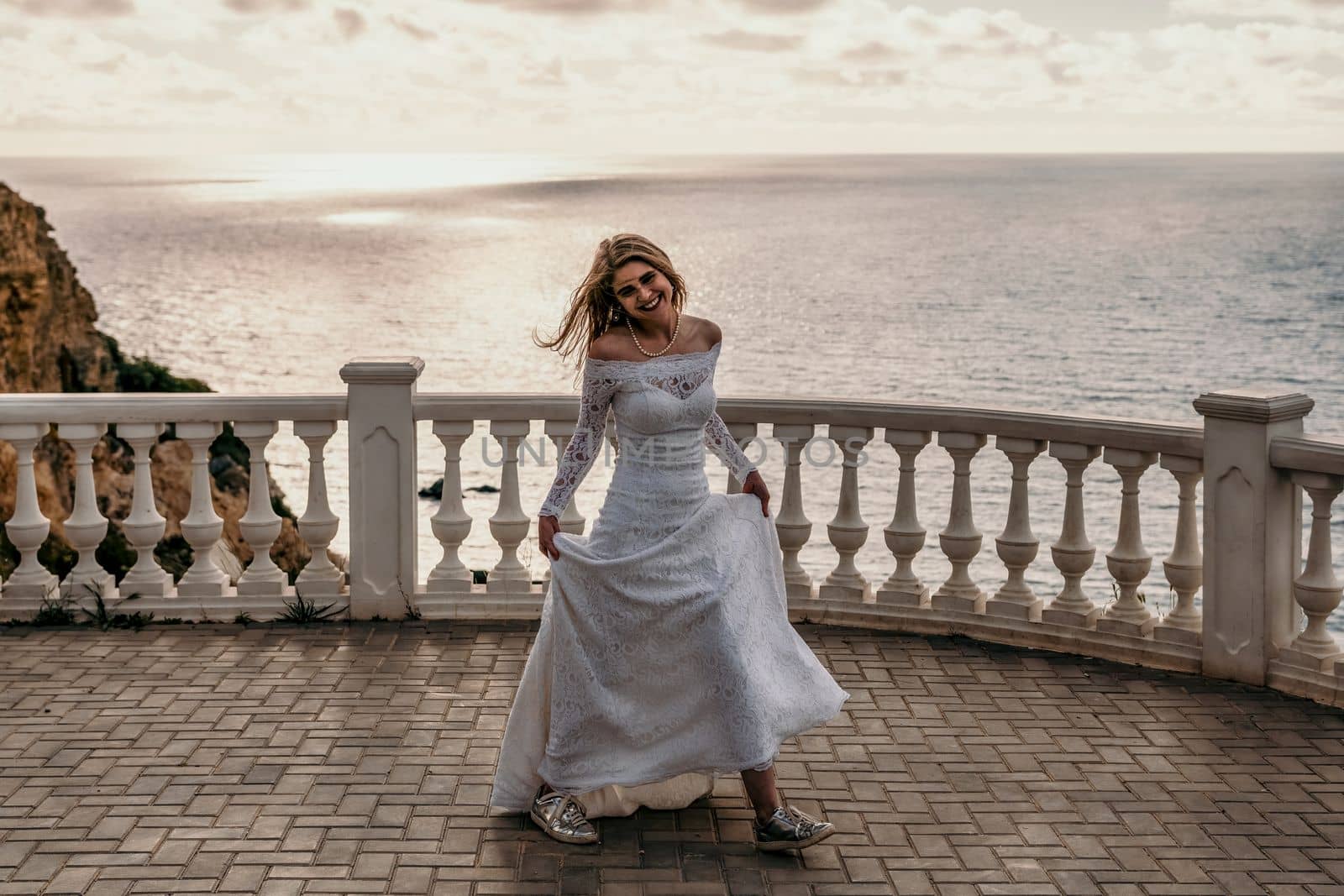 Romantic bride, blond girl in white wedding dress with open shoulders posing on open terrace with backdrop of the sea and rocks. Stylish young woman standing on terrace and looking on ocean at sunset by panophotograph