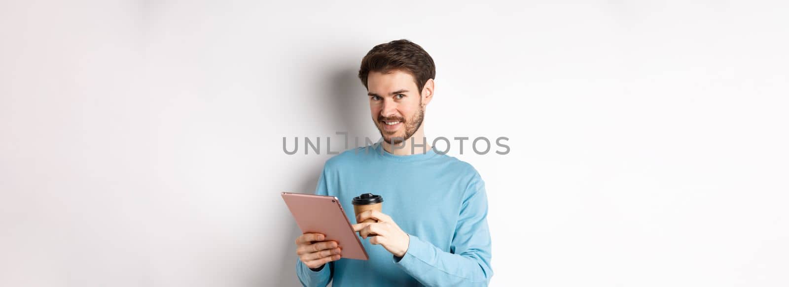 Handsome bearded man smiling at camera, drinking coffee and reading on digital tablet, standing over white background by Benzoix