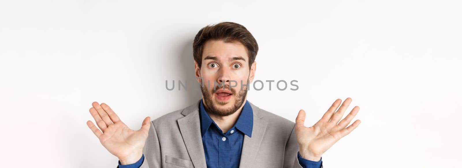 Surprised business man in suit spread hands sideways, looking confused and startled, white background.