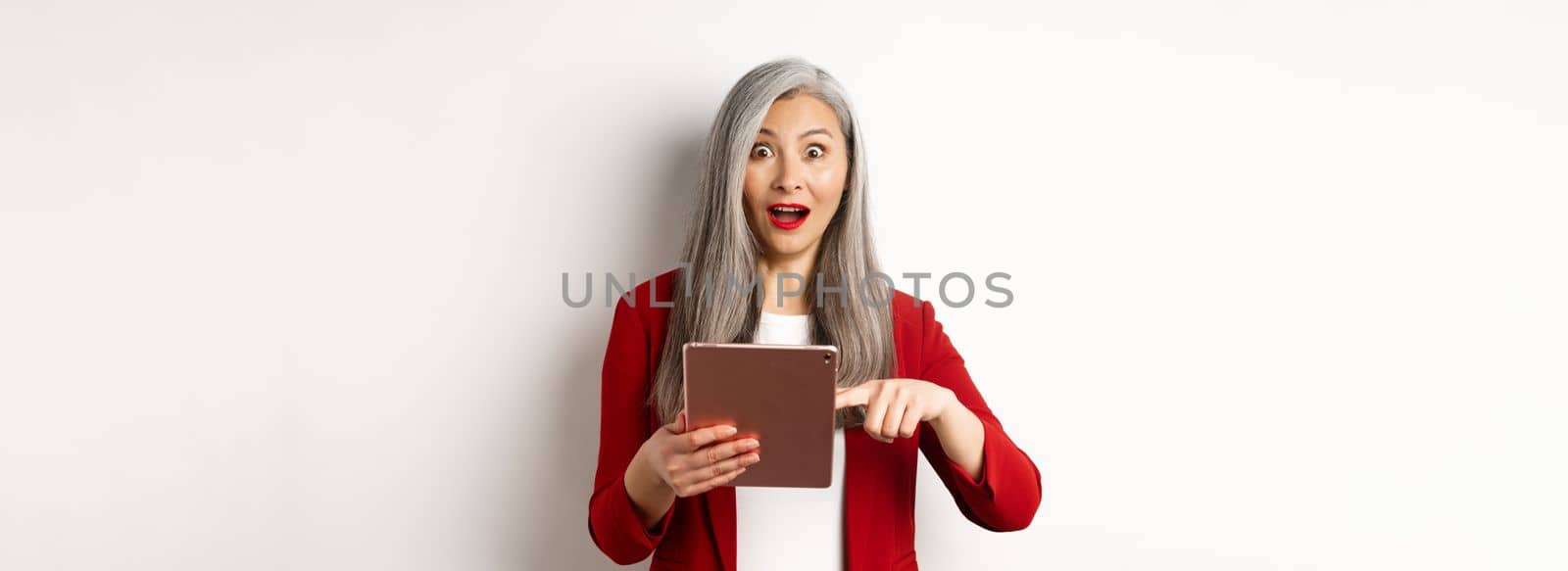 Business. Surprised female senior employer pointing at digital tablet screen, staring amazed at camera, checking out online promotion, white background.